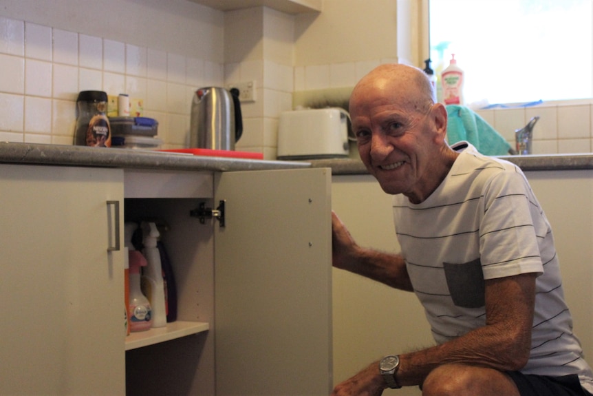 An elderly man crouches down next to a kitchen cupboard