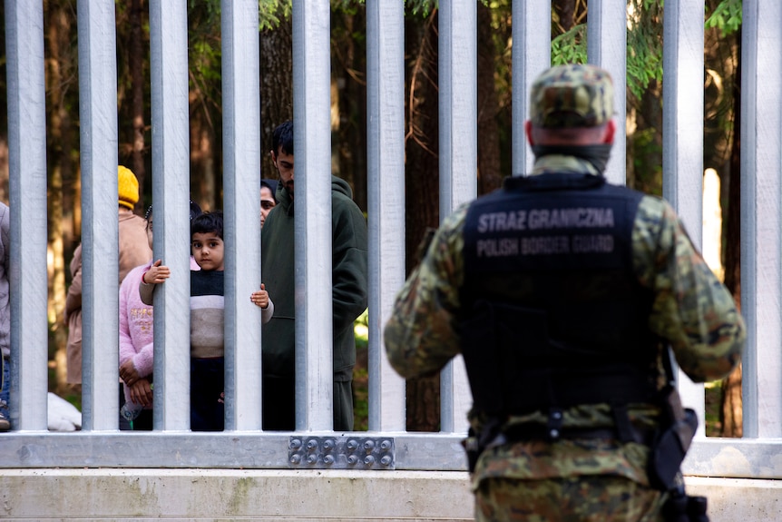 A guard is seen in the foreground while children are behind the fence in the background