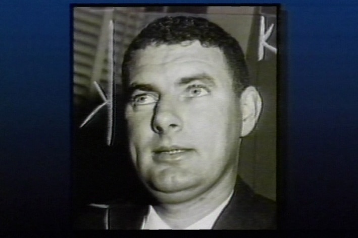 A black and white still image of a man with dark hair looking to the side of the camera.