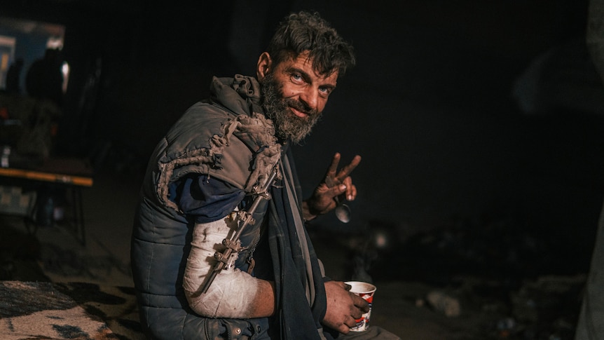 Injured man in arm sling sits with cup in hand, holding peace sign up with other hand.