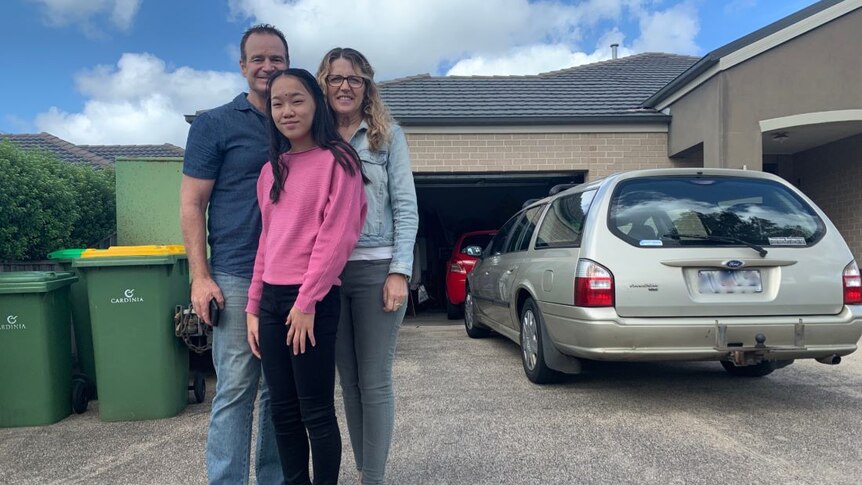 Carmel, Collin and their daughter Zhimei stand on their driveway with two cars parked behind them.