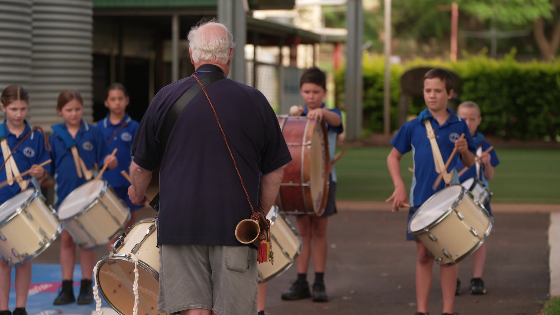 Man with his back to the camera leads a primary school-aged band of buglers and drummers.