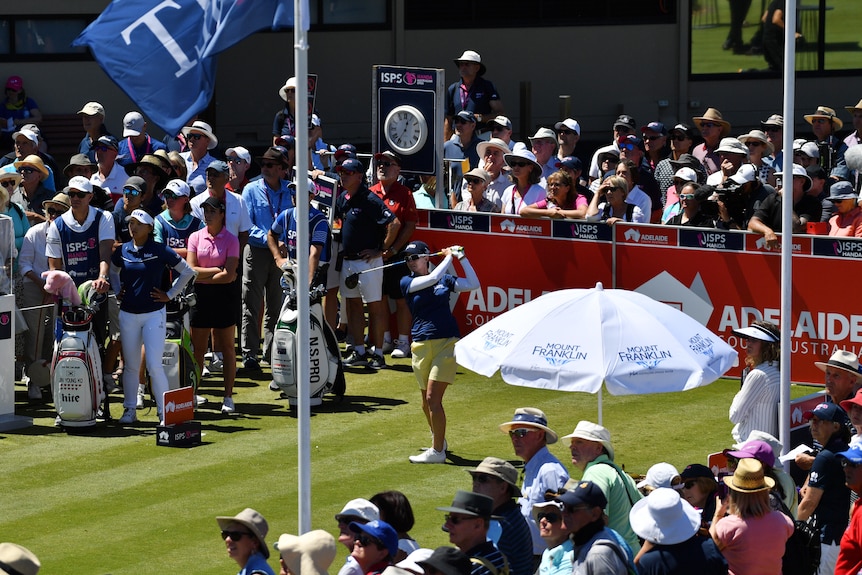 A woman golfer hits off the tee surrounded by a big crowd, with a sign behind her saying "Adelaide, South Australia".