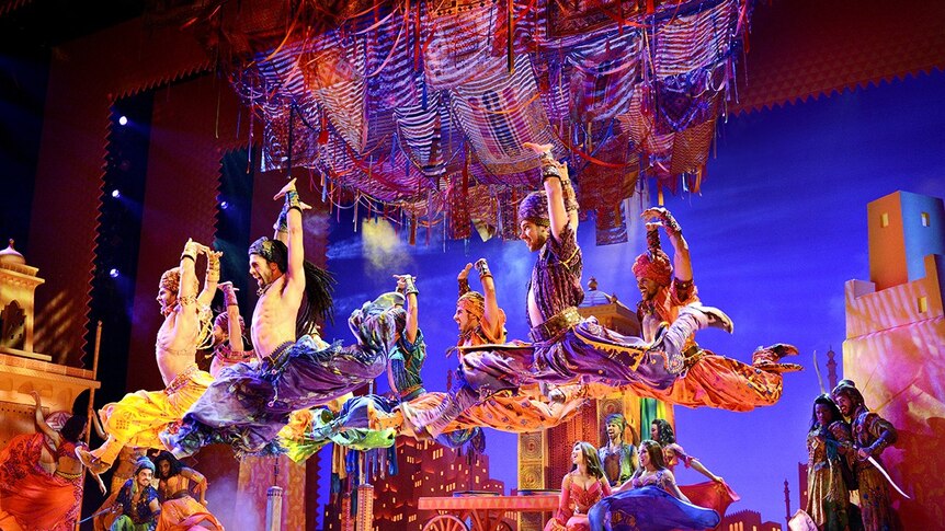 Men dressed as middle eastern sales merchants, like aladdin, perform on stage in vibrant costumes.