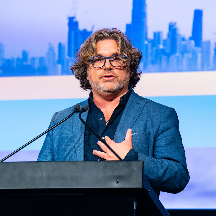 A man with glasses and wearing a suit stands behind a lectern with microphones pointed at him