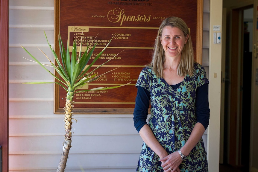 A woman with shoulder-length blonde hair stands near the front door of a weatherboard building.