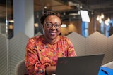 An african woman sits behind a laptop with a glass wall behind her looking at the camera 