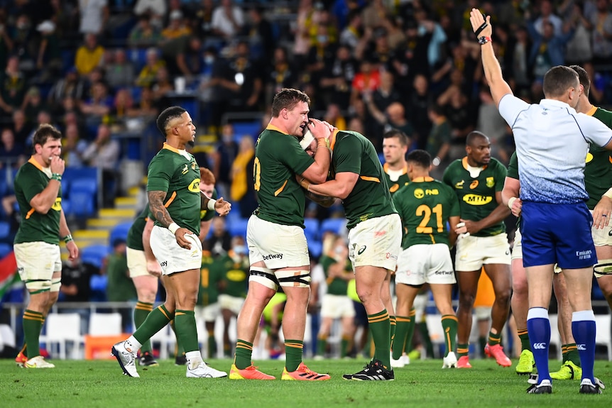 A group of Rugby players in green jerseys and white shirts hug and celebrate as a referee blows full-time in stadium