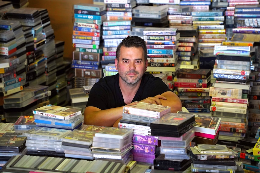 A man wearing a black t-shirt stands amongst a pile of colourful video tapes