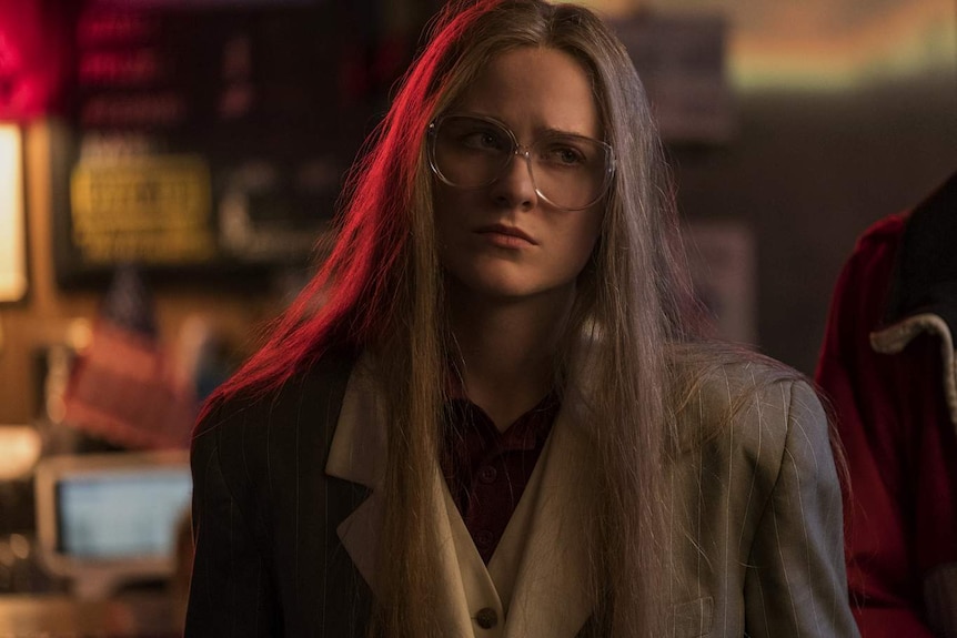 Evan Rachel Wood wearing 80s style blazer and glasses, looking concerned, in red-lit night-time interior.