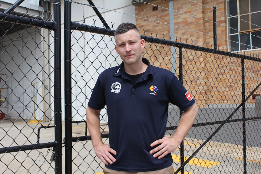 A man in a dark blue polo shirt stands with his hands on his hips in front of a locked gate.