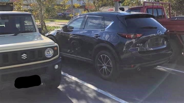 A dark-coloured Mazda 3 parked under security cameras without its numberplates. It stands between a cream Suzuki and a red ute