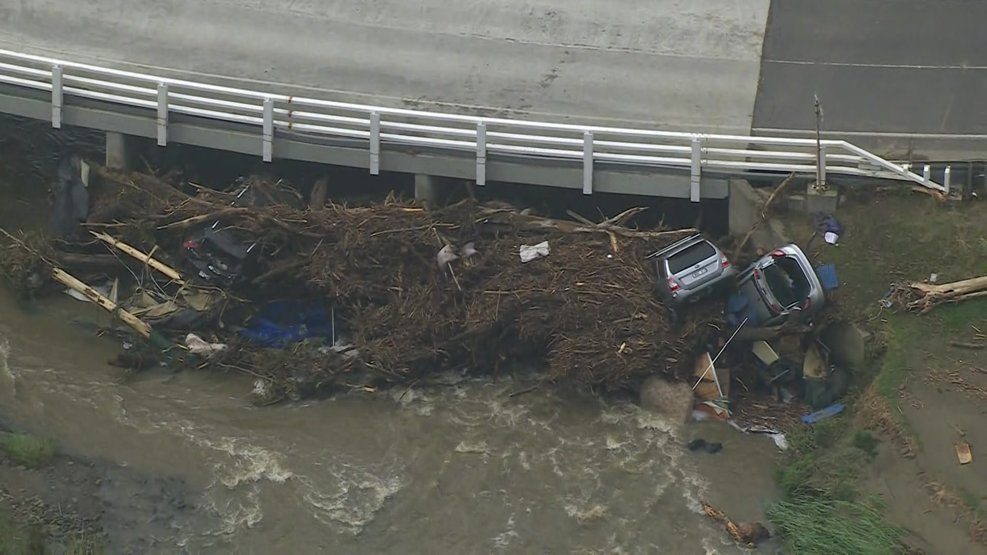 Dois carros estão meio enterrados em uma grande pilha de gravetos e detritos na água, empurrados contra uma ponte.