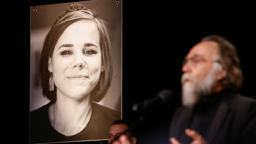 A large black and white portrait of a woman is in focus in the background as a man speaks in the foreground. 