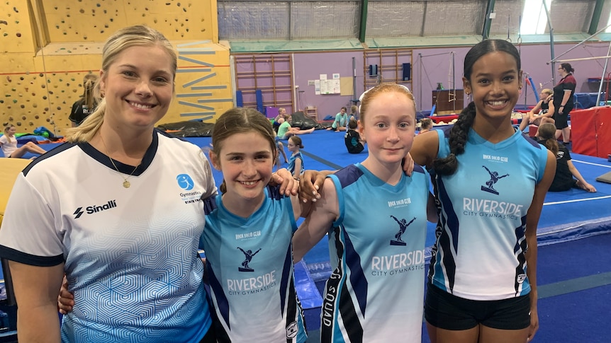 A gymnastics coach and three students stand in gymnastics clothing, in a stadium