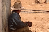 Man in an akubra squatting on ground next to a tank looking away from camera.