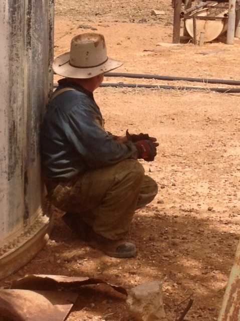 Man in an akubra squatting on ground next to a tank looking away from camera.