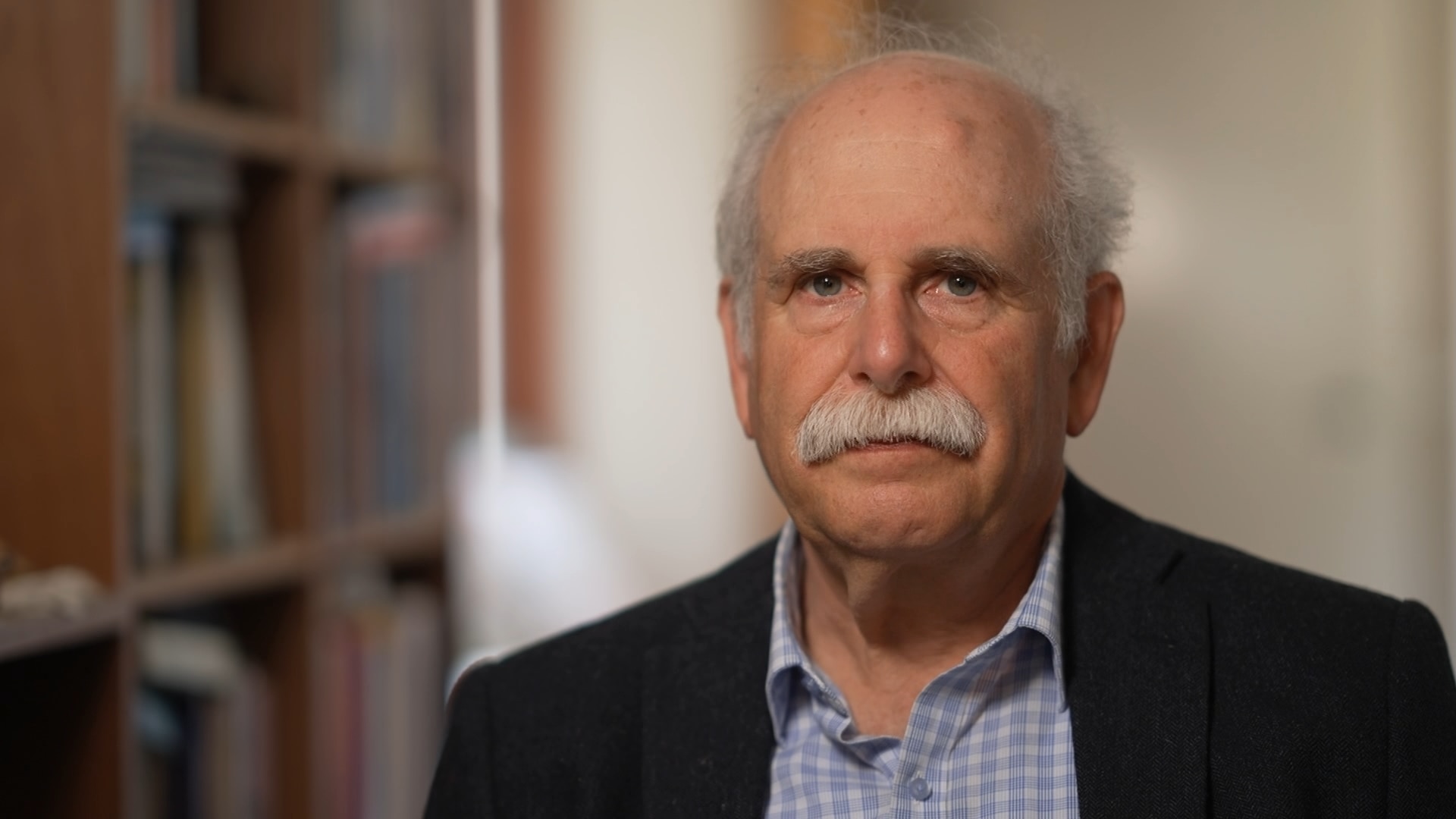 An older man with a thick grey moustache wearing a black suit jacket and blue checkered shirt sits next to bookshelves.