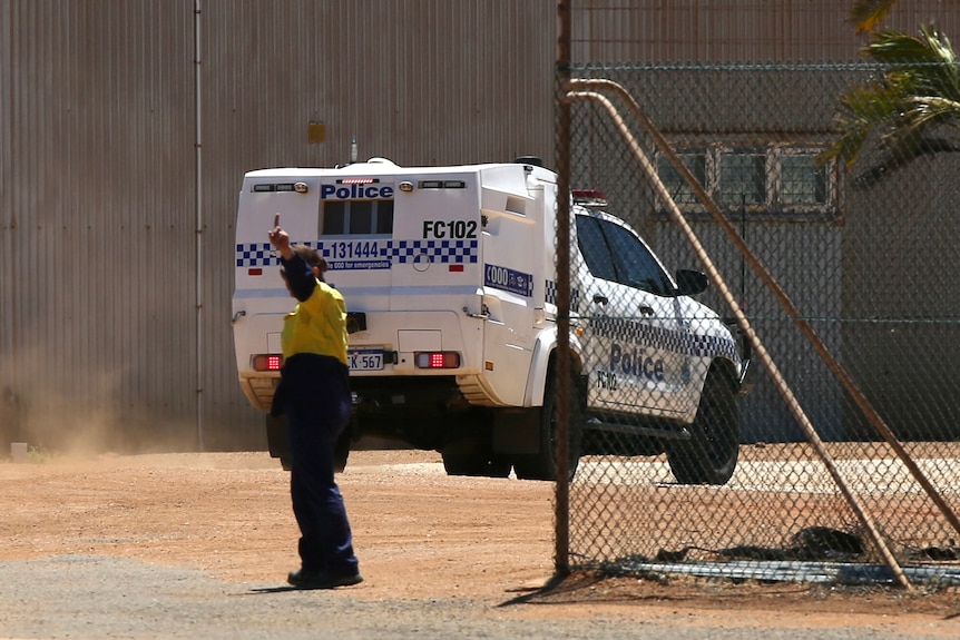 A police car drives through a gate with a man wearing hi-vis in the foreground.