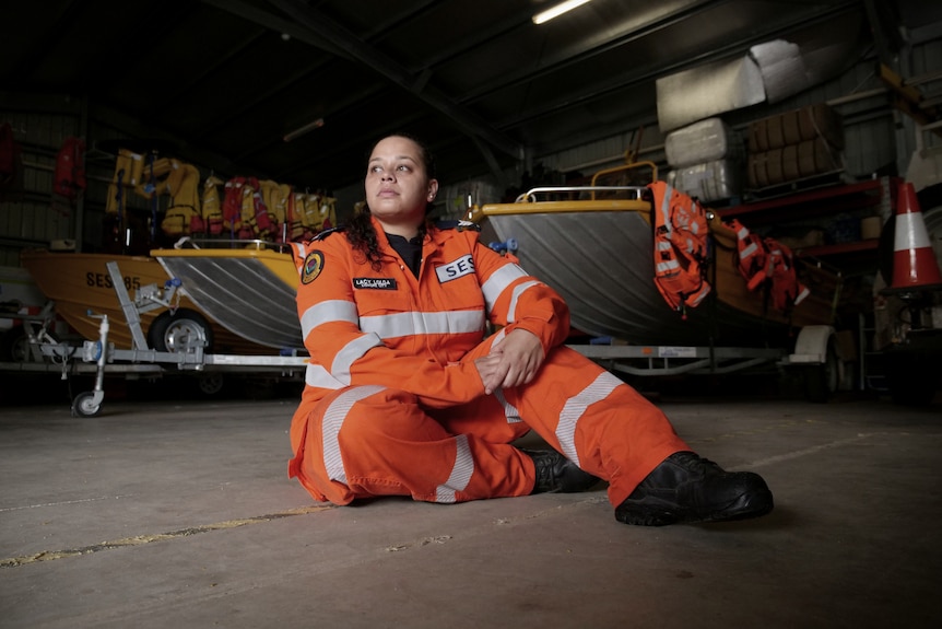 A woman in orange SES uniform sitting on the ground in a shed full of boats and SES equipment