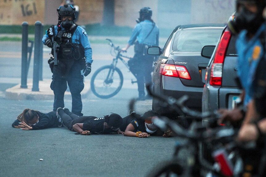 A police officer brandishing a weapon and wearing a mask looks down as three people lay face down on the ground.