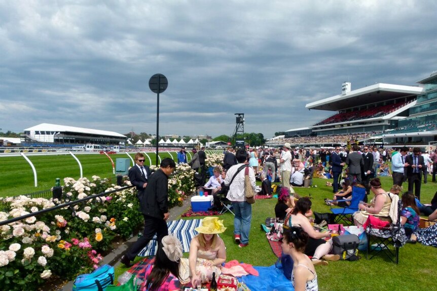 Racing fans wearing dresses and hats sit on picnic blankets beside the roses at Flemington.