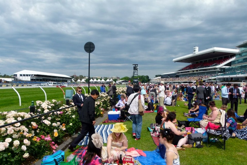 Race fans wearing dresses and hats sit on picnic rugs next to the roses in Flemington.