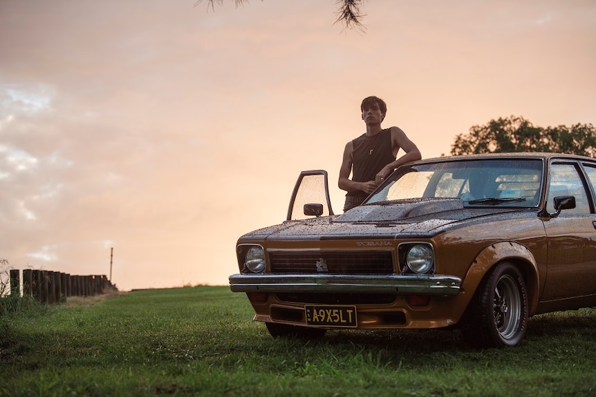 A young male stands backlit in the open door of a bronze old model Torana