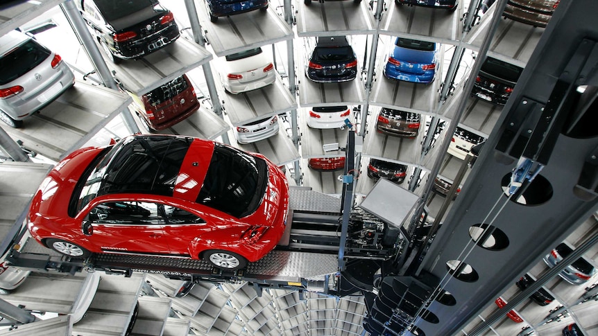 View down a deep cylindrical shaft of parked cars as a machine lifts a red VW Beetle lifted into place.
