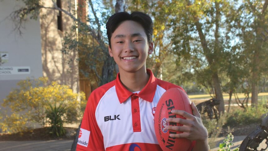 A smiling Korean boy in a red AFL uniform holding a ball.