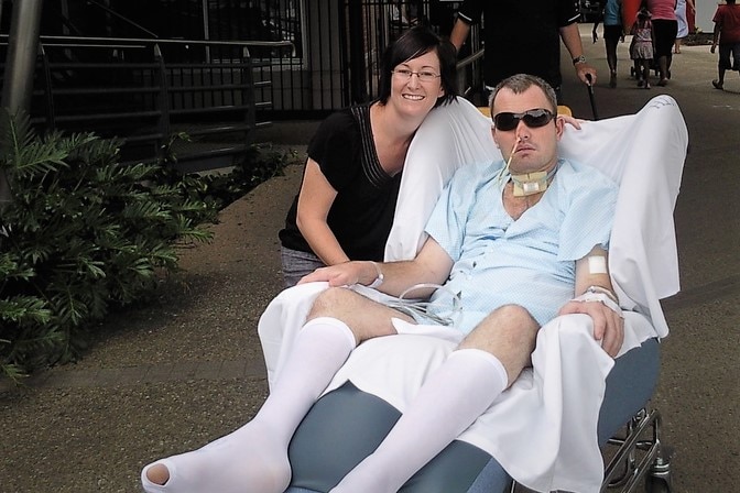 Man with sunglasses in a hospital gown, on a sofa wheelchair, with a drip connected to his nose, band aids on his arm.