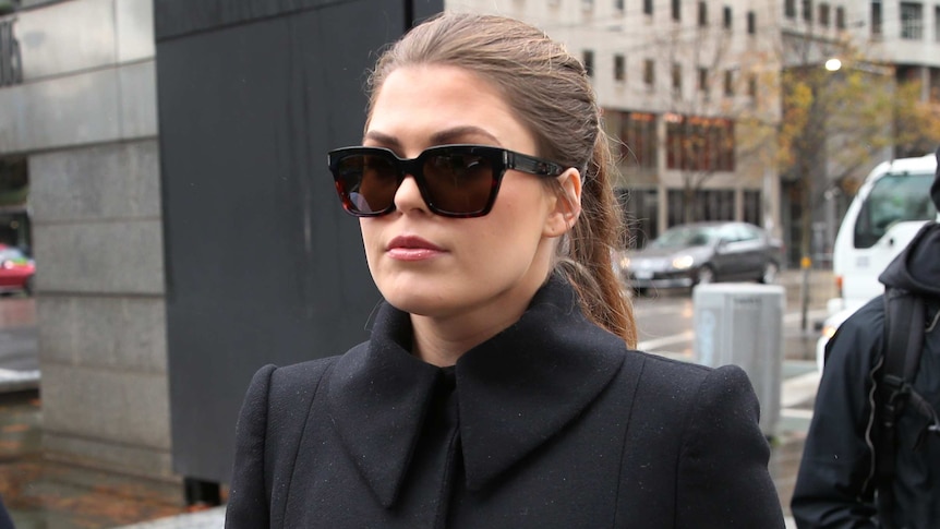 Belle Gibson, with her hair in a ponytail, wearing black sunglasses and a black coat.