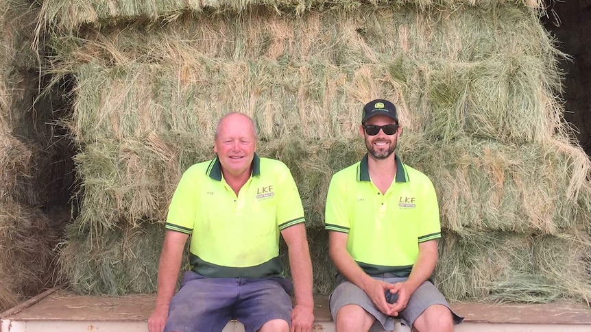 Two men in high visibility shirts sit in front of a stack of baled hay.