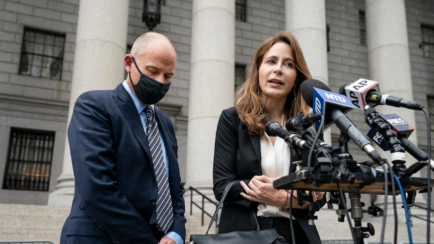 A woman speaks into microphones while a man in a navy suit and a face mask looks down.