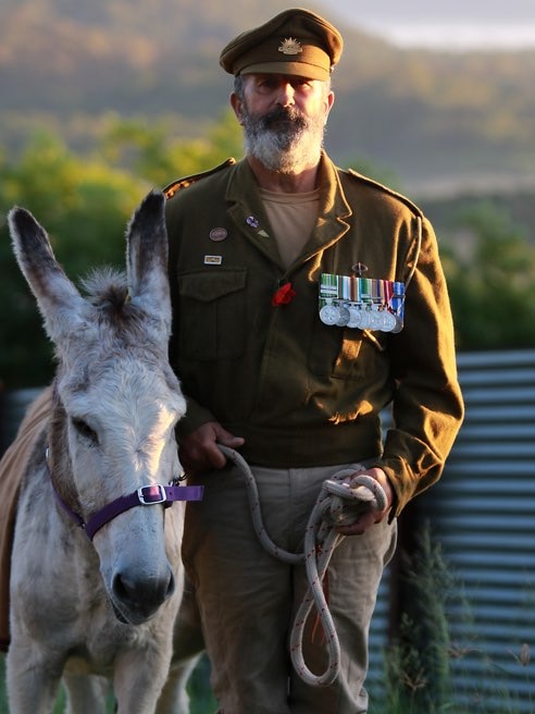 Man stands dressed in army uniform holding a lead to a grey donkey
