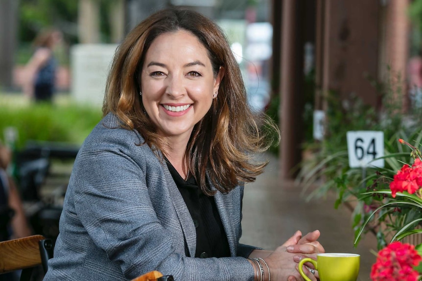 A woman wearing a grey jacket sits at a cafe, smiling.