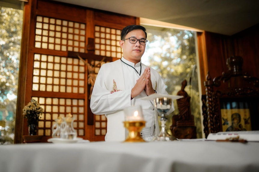 A man in priest's robes puts his hands together in prayer, in front of an altar with candles and offerings