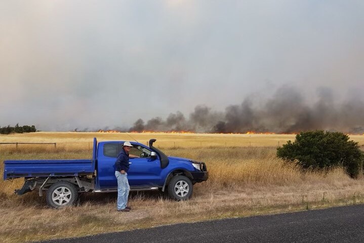 A blue ute parked with a grass fire burning behind it, in the distance