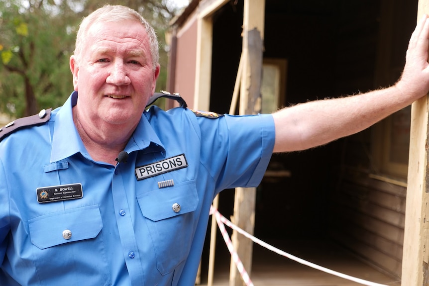 A uniformed prison officer stands next to an old forestry hut.