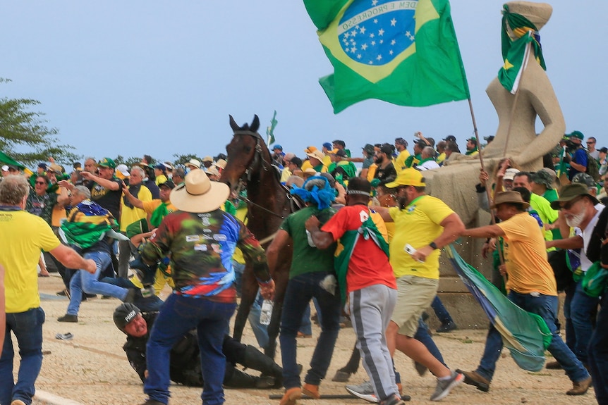 A police officer falls rom his horse as crowds wearing the brazilian flag advance on him. 