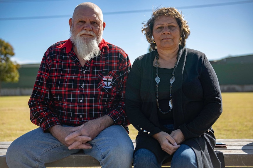 A man and a woman sit on a park bench and look at the camera.