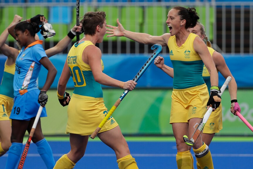 Two female hockey players wearing green and gold  joyously prepare to embrace on the hockey field.