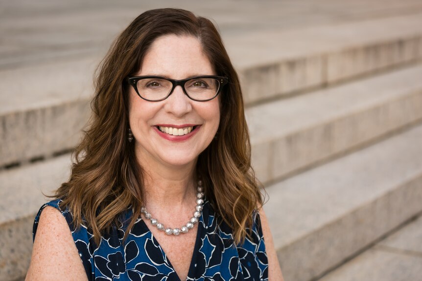 A middle-aged woman wearing a blue and black sleeveless shirt with a pearl necklace and black-rimmed glasses smiles to camera