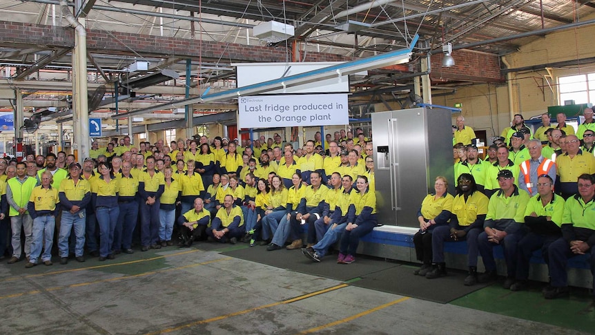 Workers in high visibility gear in a factory with the sign last fridge produced in the Orange plant