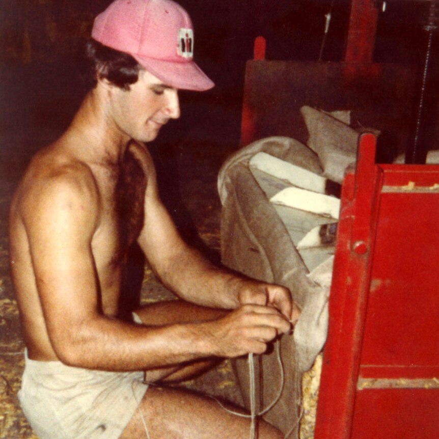 A young Italian man works on a tobacco baling machine. He's smiling while working topless, wearing a red cap and white shorts