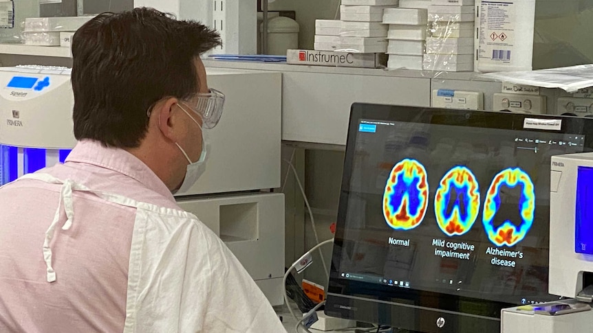 A man in medical protective gear looks at a computer with scans of brain with various degrees of dementia.