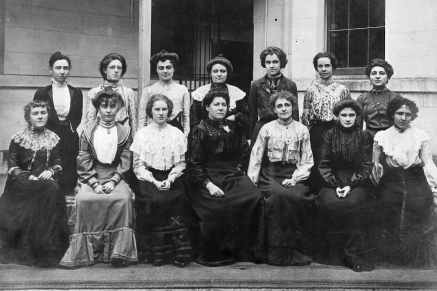 A historic black and white formal photo of 14 women in Victorian era clothing.