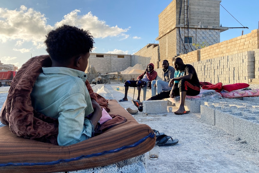 A man lies on a mattress outside a brick factory building, three more men sit opposite him