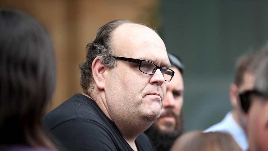 Stewart Johnston, standing in a crowd, frowns as he speaks to reporters.