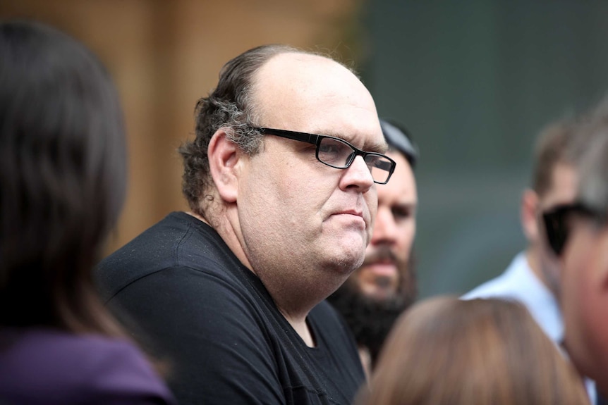 Stewart Johnston, standing in a crowd, frowns as he speaks to reporters.